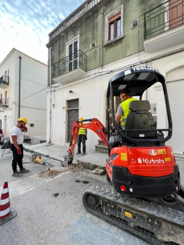Risanamento idrico a Monte Sant'Angelo: miniescavatore e operai nel cantiere stradale
